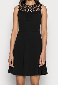 Svart ärmlös klänning med en blommig spetskrage, åtsittande bodice och utsvängd kjol som slutar ovanför knäna. Texturerad spetssmyckning.
