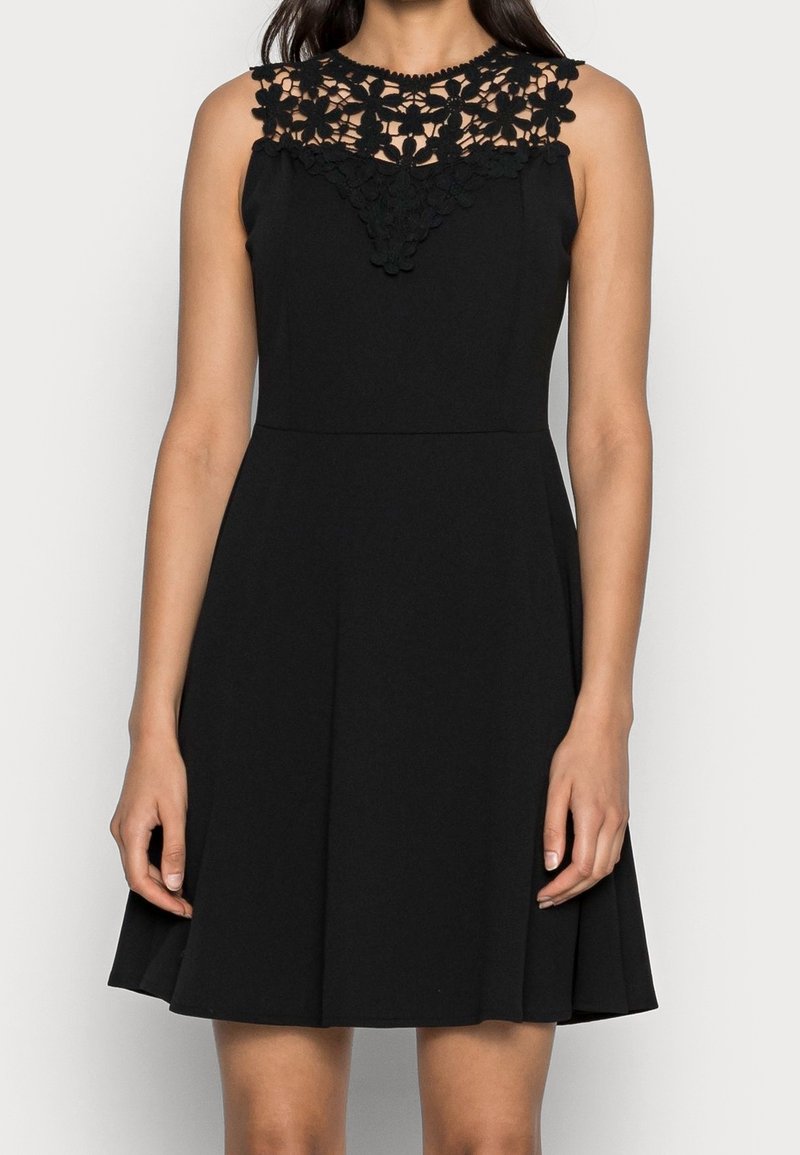 Svart ärmlös klänning med en blommig spetskrage, åtsittande bodice och utsvängd kjol som slutar ovanför knäna. Texturerad spetssmyckning.