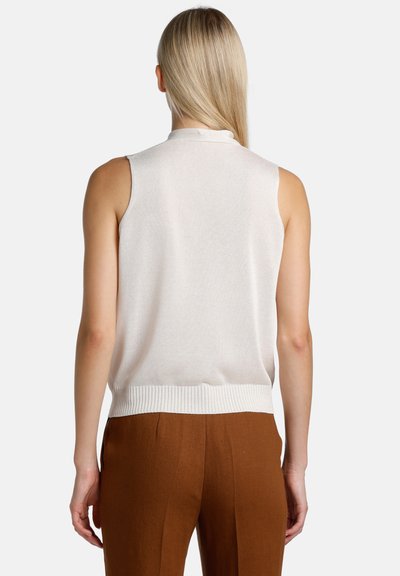 Moteris su tiesiais šviesiais plaukais, vilkinti sleeveless baltą mezginį viršų ir rudus kelnes, žiūrima iš nugaros baltame fone.