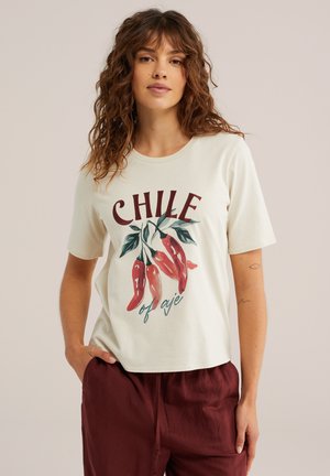 Giovane donna con capelli ricci indossa una maglietta color crema con chili rossi e la scritta "CHILE of aje", abbinata a pantaloni color vinaccia, sfondo neutro.