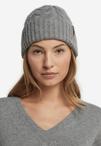 Grå strikket beanie med en tekstureret mønster og ribbet design, med et lille lærremsaccent på siden. Ideel til brug i koldt vejr.