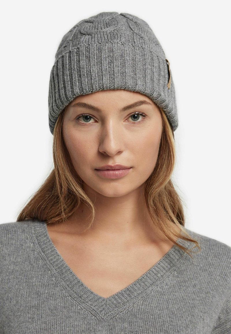 Grå strikket beanie med en tekstureret mønster og ribbet design, med et lille lærremsaccent på siden. Ideel til brug i koldt vejr.