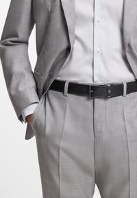 Completo grigio chiaro con finitura strutturata, composto da blazer monopetto, pantaloni plissettati e cintura in pelle nera con fibbia metallica.