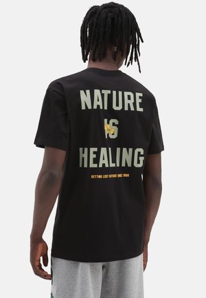 Mann mit Dreadlocks trägt ein schwarzes T-Shirt mit dem Schriftzug „Nature is Healing“ und einer gelben Blumen-Grafik auf dem Rücken, kombiniert mit grauen Shorts.
