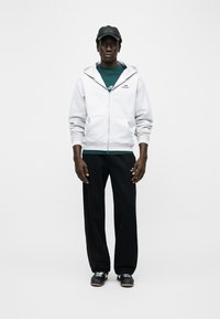 Άνδρας που φοράει μαύρο καπέλο, ανοιχτό γκρι ζακέτα με φερμουάρ πάνω από πράσινο πουκάμισο, μαύρη φόρμα και μαύρα παπούτσια, στέκεται μπροστά σε λευκό φόντο.