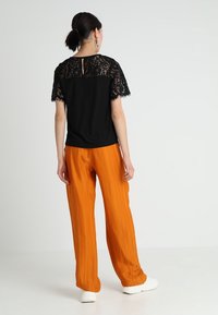 Top negro con mangas de encaje y un detalle de ojal en la parte trasera, combinado con pantalones naranjas plisados y zapatillas blancas, sobre un fondo neutro.