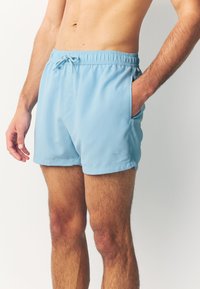 Shorts de bain bleu clair en tissu lisse et léger. Ils sont dotés d'une taille élastique avec un cordon de serrage et de poches latérales.