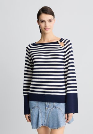 Maglione a righe blu navy e crema con scollatura ampia e un dettaglio a bottone. Abbinato a una gonna di jeans con fondo svasato e cuciture in vista.
