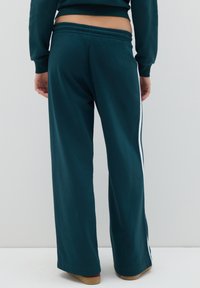 Persona vestida con pantalones holgados de color teal oscuro con franjas blancas a los lados y un sudadera corta a juego, de pie frente a una pared blanca lisa.