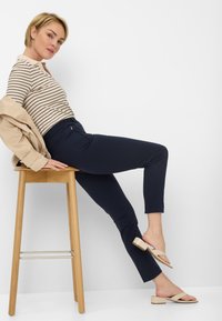 Marineblaue Hose mit einer figurnahen Silhouette und einem strukturierten Bund. Das Model trägt ein gestreiftes Langarmoberteil und hellfarbige Absatzsandalen.