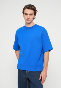 Νεαρός άνδρας με κοντά καστανά μαλλιά, φοράει ένα απλό φωτεινό μπλε φαρδύ T-shirt και σκούρο μπλε τζιν, στέκεται μπροστά σε λευκό φόντο.