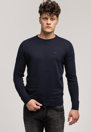 Granatowy sweter dzianinowy z okrągłym dekoltem, ściągaczami na mankietach i u dołu, z małym logo na piersi. Gładka faktura, dopasowany krój.