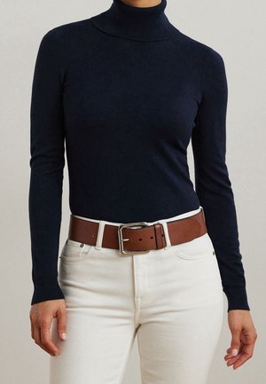 Personne portant un pull à col roulé bleu marine rentré dans un pantalon blanc avec une large ceinture en cuir marron et une boucle argentée.