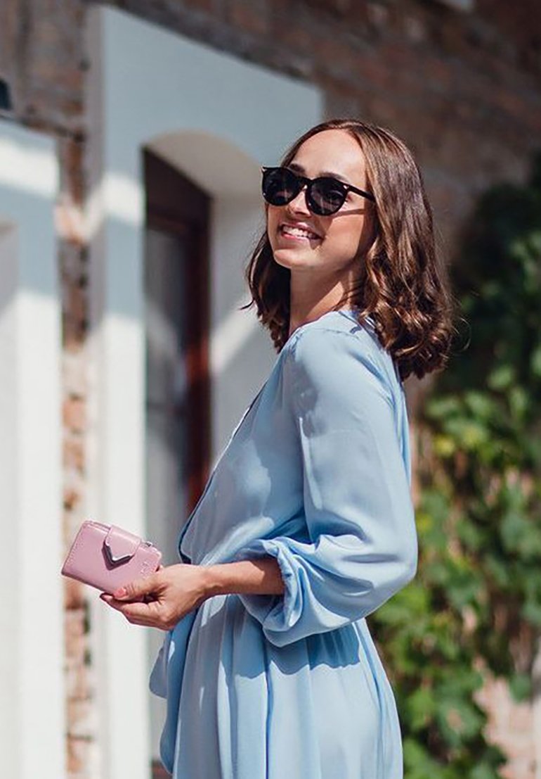 Light blue dress with long, billowy sleeves. Black sunglasses. Holding a small, pink wallet with a clasp detail. Vintage brick wall backdrop.