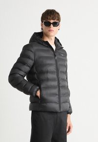 Giubbotto piumino nero con cappuccio, caratterizzato da un design trapuntato e una chiusura frontale con zip. La finitura testurizzata conferisce calore e un aspetto elegante.