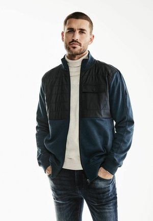 Chaqueta de navy con cremallera y yugo acolchado negro, cuello alto y bolsillo en el pecho. Lleva puesta una camiseta de cuello alto clara y se combina con jeans oscuros.
