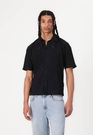 Camisetta nera a maniche corte con texture a righe verticali, apertura con bottoni e vestibilità ampia; abbinata a jeans azzurri chiari, a taglio largo.