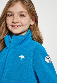 Giacca in pile blu brillante con zip, colletto alto e texture morbida. Presenta un logo a forma di ombrello bianco e una patch rotonda sulla manica.