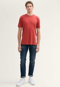 Κόκκινο βαμβακερό μπλουζάκι με κοντά μανίκια, στρογγυλή λαιμόκοψη και εκτυπωμένο σχέδιο στην αριστερή πλευρά. Συνδυασμένο με σκούρο μπλε τζιν και λευκά αθλητικά παπούτσια.