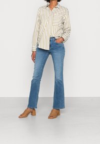 Chemise boutonnée beige et blanche à rayures, jeans évasés bleus, et chaussures à talons ouvertes couleur beige. Comprend une poche plaquée sur la chemise.