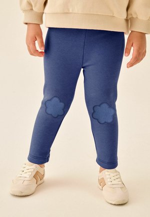 Kind draagt blauwe leggings met bloemenpatches op de knieën en witte sneakers met beige details, staand op een effen achtergrond.