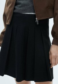 Jupe noire plissée en tissu à texture lisse, avec une ceinture. Associée à un pull gris et une veste aviateur marron.