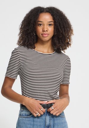 Jeune femme aux cheveux bouclés portant un haut à manches courtes rayé noir et blanc et un jean bleu, ajustant sa ceinture noire.