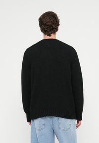 Czarny sweter z dzianiny, luźny krój, teksturowany materiał, dekolt w kształcie V na plecach, ściągacze przy mankietach i u dołu, zestawiony z jasnoniebieskimi dżinsami.