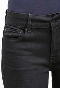 Jeans ajustados negros con una textura suave, tiro alto y un ligero desgastado. Presenta herrajes en tono cobre y un pequeño logo en el bolsillo frontal.