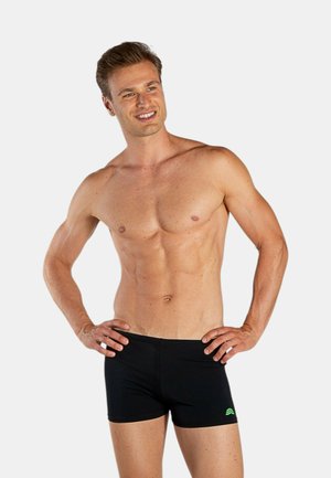 Uomo sorridente a torso nudo in piedi con le mani sui fianchi, indossa pantaloncini da bagno neri con un piccolo logo verde, su sfondo chiaro e uniforme.