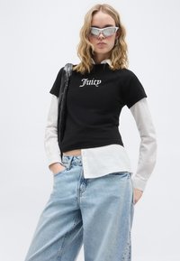 Γυναίκα που φοράει ασημένια γυαλιά ηλίου σε σχήμα γάτας, με στρωτό μαύρο μπλουζάκι "Juicy" πάνω από άσπρο πουκάμισο, ανοιχτόχρωμο μπλε τζην και κρατώντας μαύρη τσάντα ώμου.