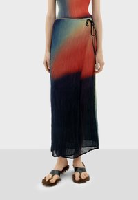 Plooien maxi jurk in een verloop van rood, blauw en groen. Heeft een strik tailleband en een transparante textuur. Gecombineerd met zwarte sandalen met bontaccenten.