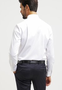 Biele košeľové tričko s klasickým golierom a dlhými rukávmi. Spárované s tmavomodrými nohavicami so čiernym opaskom a decentnými zadnými vreckami.