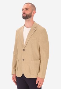 Blazer in lino beige con finitura testurizzata, due tasche frontali, tre bottoni e una tasca sul petto. Indossato sopra una camicia bianca.