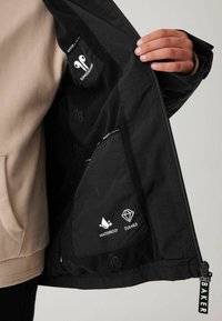 Die Innenausstattung der schwarzen wasserfesten Jacke verfügt über Taschen mit den Logos "Langlebig" und "Wasserfest", strukturierter Stoff und einen diagonalen Reißverschluss.