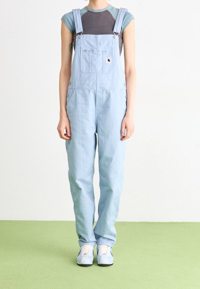 Helleblaues Denim-Overall mit verstellbaren Trägern, zwei seitlichen Taschen und einer Brusttasche; getragen über einem kurzärmeligen grauen Oberteil und hellen Schuhen.