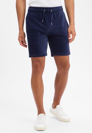 Männliches Model trägt marineblaue, gerippte Shorts mit Kordelzug und weiße Sneaker, steht vor einem schlichten weißen Hintergrund.