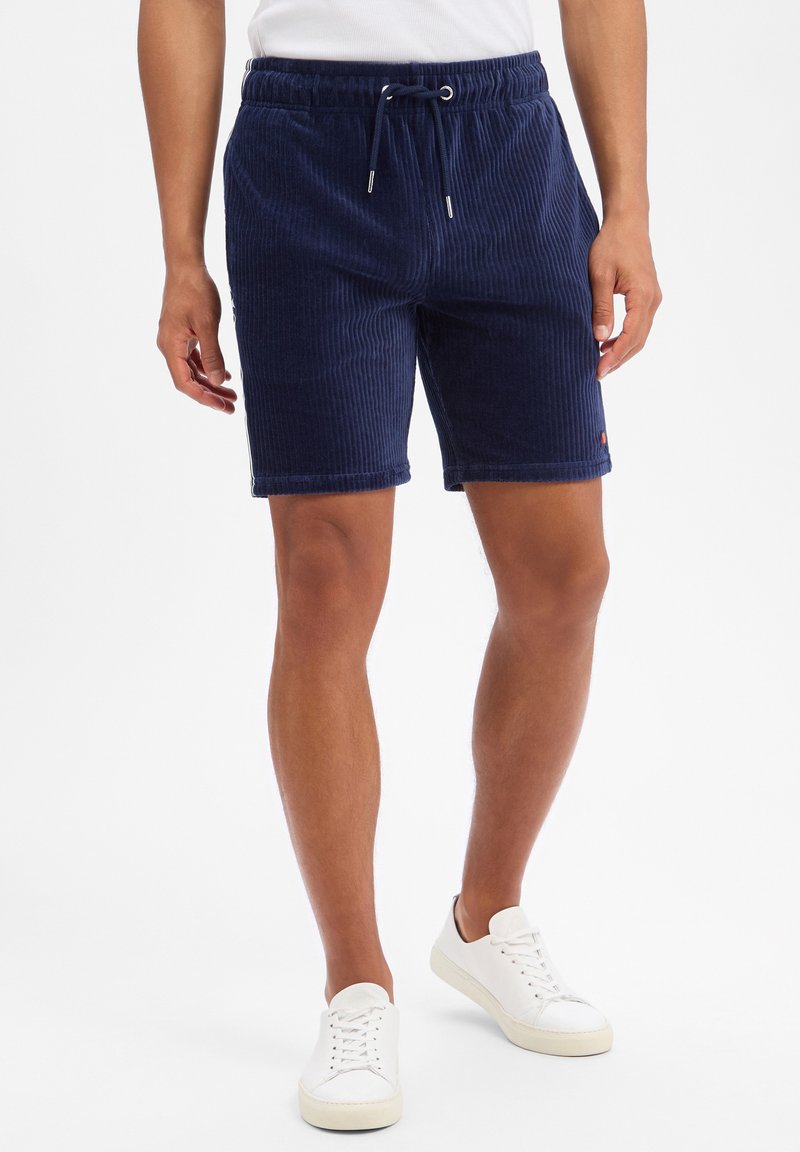 Male model wearing navy blue ribbed drawstring shorts and white sneakers, standing against a plain white background.