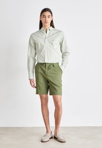 Cămașă cu mânecă lungă în carouri verzi și albe, pantaloni scurți verzi cu bentiță ajustabilă, și mocasini gri. O ținută simplă, casual, cu linii clare.