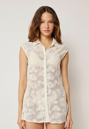 Frau mit schulterlangen braunen Haaren, die eine ärmellose, geknöpfte, cremefarbene, transparente Bluse mit floralem Spitzenmuster trägt, vor einem einfarbigen Hintergrund stehend.