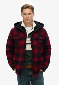 Rød og marineblå ternet flannel skjorte med hætte, der har knappede brystlommer og et lagdelt look over en grøn og hvid stribet skjorte.