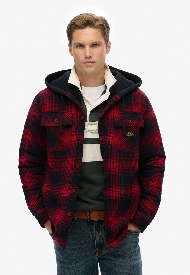 Rød og marineblå ternet flannel skjorte med hætte, der har knappede brystlommer og et lagdelt look over en grøn og hvid stribet skjorte.