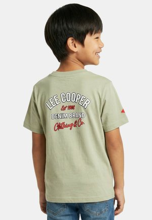 Garçon souriant regardant vers la droite, portant un t-shirt vert clair Lee Cooper avec un logo rouge et blanc au dos, et un jean bleu.
