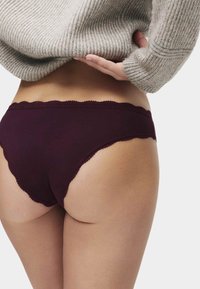Burgundské bavlněné spodní prádlo s vlnkovým krajkovým lemem, které má nízký pas a jemnou strukturu, částečně zakryté šedým svetrem.
