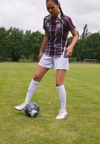 Maillot de football rayé violet et blanc, short blanc et chaussettes blanches. Se tient sur l'herbe à côté d'un ballon de football noir et blanc.