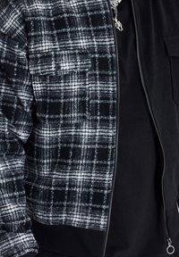 Veste zippée à carreaux noir et blanc portée sur un t-shirt noir uni avec un collier pendentif en argent.