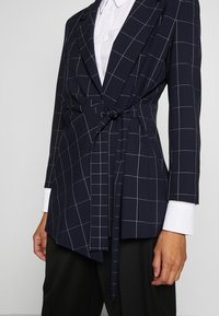 Marinblå blazer med vitt rutmönster, omslagdesign och knyt vid midjan. Har långa ärmar och vita skjortärmar som syns under.