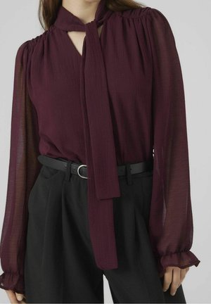 Frau mit transparenter, langärmeliger, bordeauxroter Bluse mit Schleife am Hals und schwarzen, hoch taillierten Hosen mit schwarzem Gürtel und silberner Schnalle.