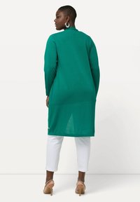 Zielony kardigan z długim rękawem o gładkiej teksturze, z okrągłym kołnierzem i bocznymi rozcięciami, noszony na białych spodniach i beżowych szpilkach.