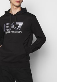 Felpa con cappuccio in cotone nero con tasca frontale, cappuccio con coulisse e logo EA7 Emporio Armani bianco con accenti lineari.
